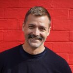 Press photo of Patrick Grant stood in front of a red wall