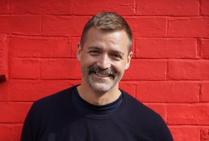 Press photo of Patrick Grant stood in front of a red wall